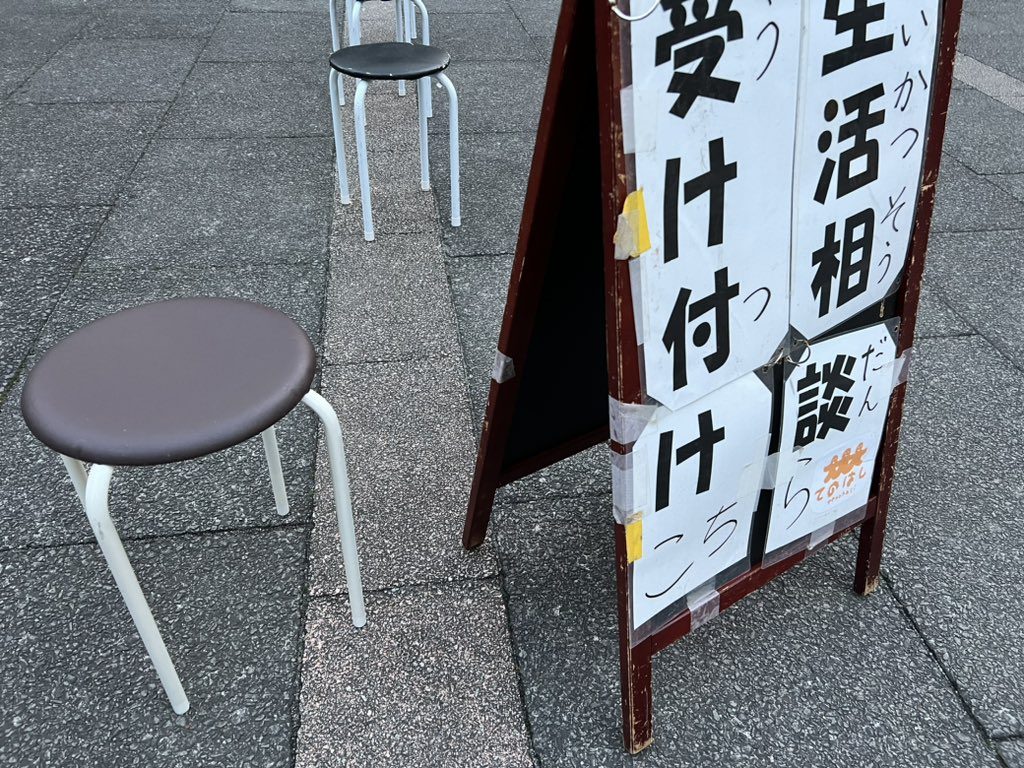 「生活相談 受け付けこちら」のA型看板と丸椅子