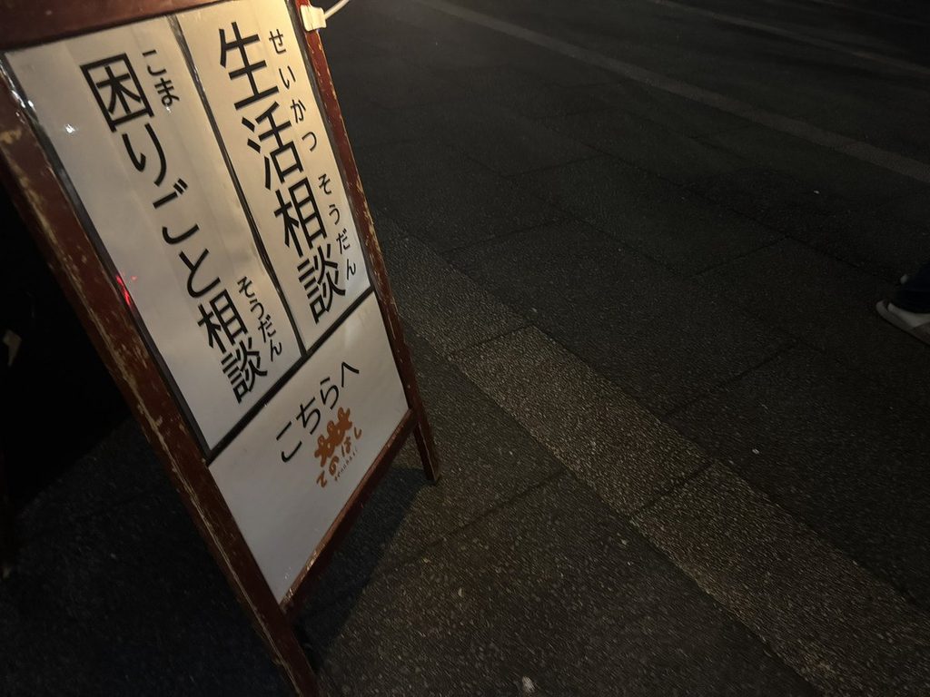 夜の会場に立つ「生活相談 困りごと相談 こちらへ」のA型看板
