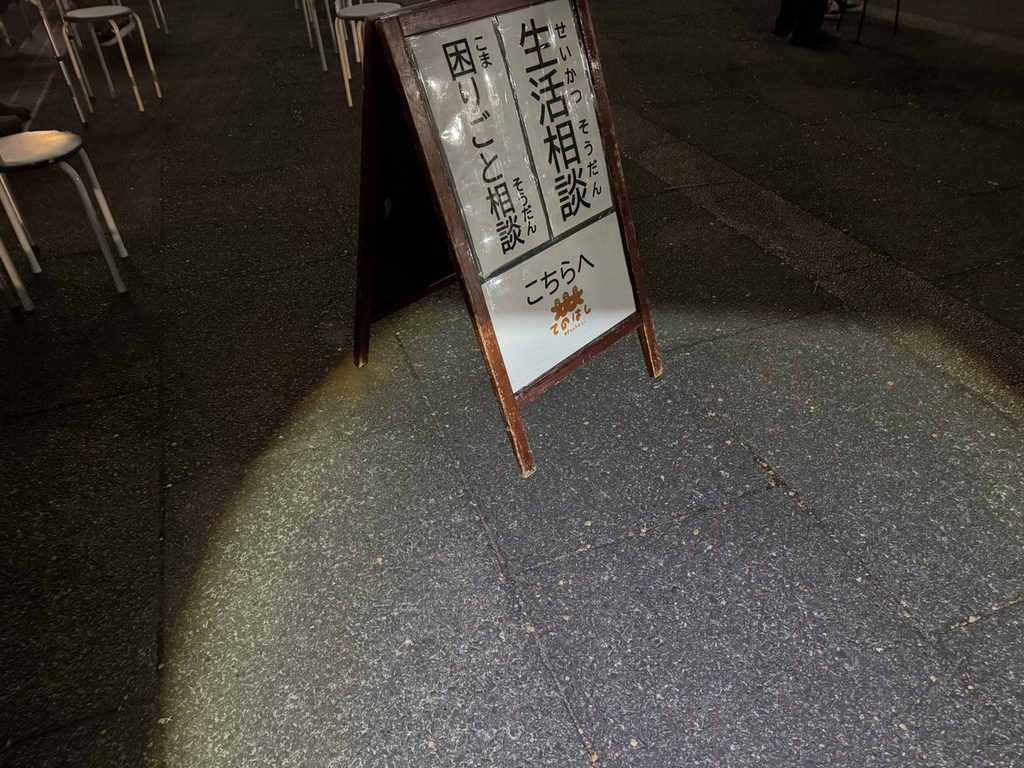 夜の会場に立つ「生活相談 困りごと相談 こちらへ」のA型看板