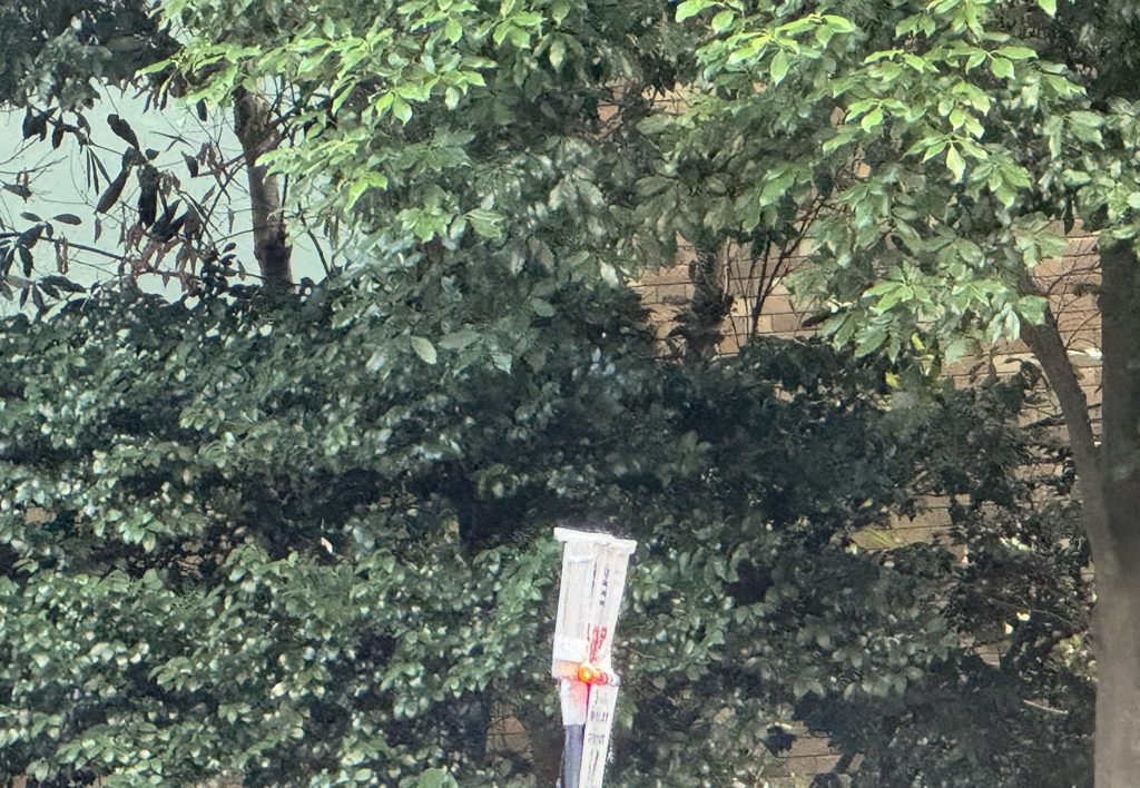 公園の緑の茂みとレンガの壁。手前に手書きの案内が掲げられている